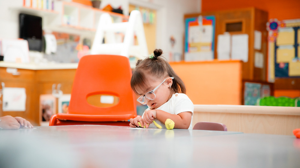 A very young child with glasses plays with play-doh at an early education center