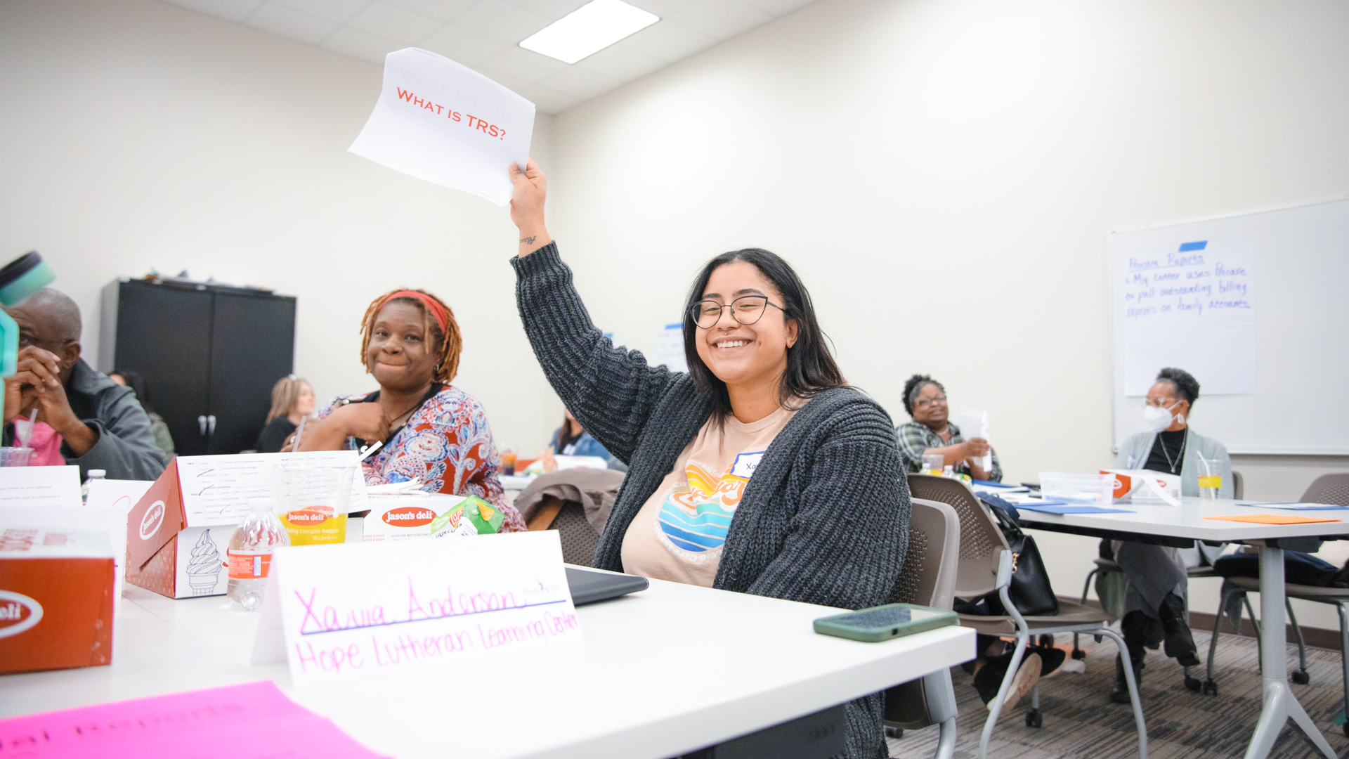 An educator at a workshop smiles and holds up a piece of paper that reads, "What is TRS?"