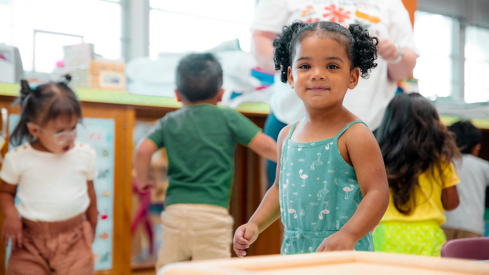 A young child in a classroom smiles at the camera